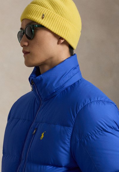 Modra puhovka z navpičnimi šivi in rumenim vezenim logotipom, kombinirana z rumeno rebrasto pleteno kapa in zelenimi sončnimi očali.