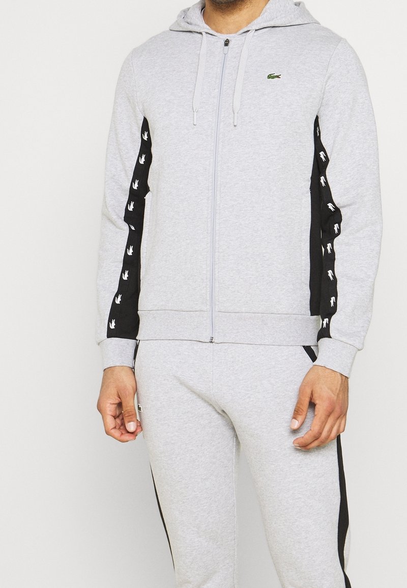 Homme portant un sweat à capuche zippé gris clair et un jogging assorti avec des panneaux latéraux noirs ornés de petits logos blancs.