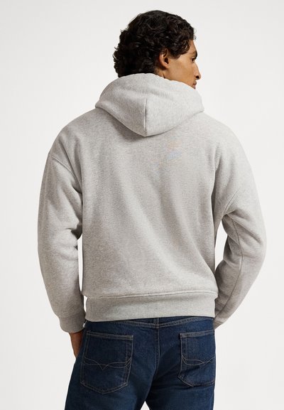 Mężczyzna o kręconych ciemnych włosach, ubrany w jasnoszary kaptur i ciemnoniebieskie dżinsy, stoi tyłem, z rękami w kieszeniach.