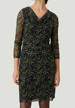 Femme portant une robe noire longueur genou avec des motifs floraux abstraits verts et des manches trois-quarts transparentes, debout devant un fond uni.
