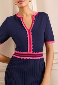 Robe tricotée bleu marine à manches courtes, avec un bord festonné rose le long du décolleté et de la taille, et une texture plissée sur la jupe.