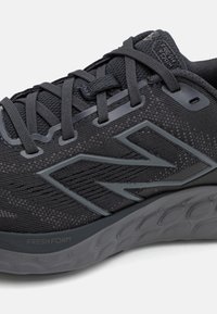 Černá sportovní bota s prodyšným síťovaným svrškem a šedými akcenty. Obsahuje flexibilní gumovou podrážku a výrazné logo "N" na boku.