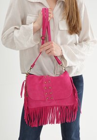 Pink suede crossbody taske med frynser, nitter og klaplukning. Den har en lang, justerbar rem for alsidighed.
