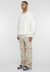 Cremefarvet sweatshirt med en lomme foran og et diskret logo, parret med beige cargo bukser med flere lommer og en løs pasform, suppleret med sorte sneakers.
