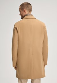 Cappotto di lana color sabbia con design dritto, revers a punte e una fessura centrale sul retro, indossato sopra un indumento marrone. Texture liscia, vestibilità su misura.