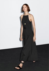 Černé halter maxi šaty mají splývavou siluetu. Doplňky zahrnují dlouhý černý náhrdelník s třásněmi. Modelka má na sobě černé sandály.