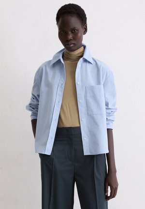 Donna nera con capelli corti che indossa un dolcevita beige, una camicia celeste con bottoni e pantaloni grigio scuro davanti a una parete bianca liscia.