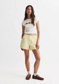Weißes, kurz geschnittenes T-Shirt mit bunten Patches auf der Vorderseite, kombiniert mit gelb gestreiften Shorts. Die Shorts haben einen elastischen Bund. Braune Sneakers.