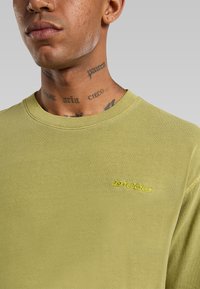 Muž s tetováním na krku, který má na sobě světle zelený svetr s kulatým výstřihem a žlutým vyšitým logem "Dickies" na hrudi.