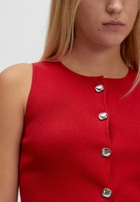 Rotes ärmelloses Oberteil mit gerippter Textur, versehen mit fünf glänzenden Metallknöpfen auf der Vorderseite. Schlichter runder Halsausschnitt ohne zusätzliche Muster.
