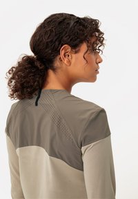 Haut de sport léger à manches longues en beige et vert olive. Présente un design perforé sur le haut du dos et une section inférieure texturée.