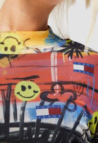 Top gráfico colorido con textura de malla, con patrones de caras sonrientes, flores y el logo "TOMMY JEANS" en acentos azules y rojos.