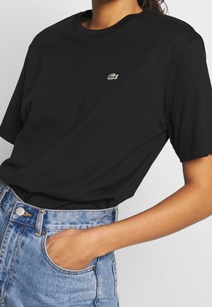 Person trägt ein schwarzes Lacoste-T-Shirt und blaue, hoch taillierte Jeans, mit einer Hand in der vorderen Tasche der Jeans.