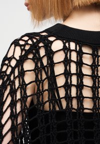 Μαύρο πλεκτό τοπ με ανοιχτό, δίχτυ σχέδιο και απλό μαύρο στρώμα βάσης, με ριμπ λαιμόκοψη. Φαίνεται εκτεθειμένο δέρμα και τιράντες.