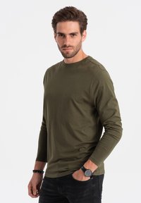 Olivegrünes T-Shirt mit langen Ärmeln, aus einem weichen Stoff, mit Rundhalsausschnitt und taillierter Silhouette, getragen zu schwarzen Jeans.