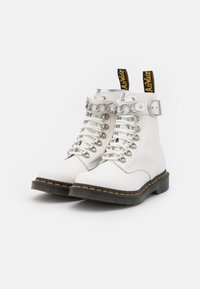 Białe skórzane botki za kostkę z ozdobnym srebrnym łańcuchem, metalowymi oczkami i białymi sznurowadłami. Czarna podeszwa z żółtym przeszyciem; charakterystyczna metka na pięcie.