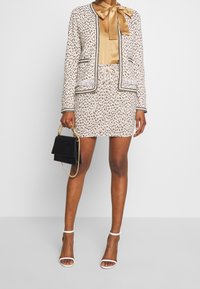 Femme portant un ensemble jupe et blazer à motifs beige, blouse en satin doré avec nœud, escarpins blancs, tenant un sac à main noir avec une anse en chaîne dorée.