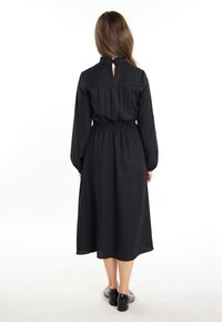 Rochie neagră cu mâneci lungi, talie elastică și o deschidere în formă de inimă în partea din spate. Țesătură fină cu o siluetă fluidă care ajunge până la jumătatea gambei.