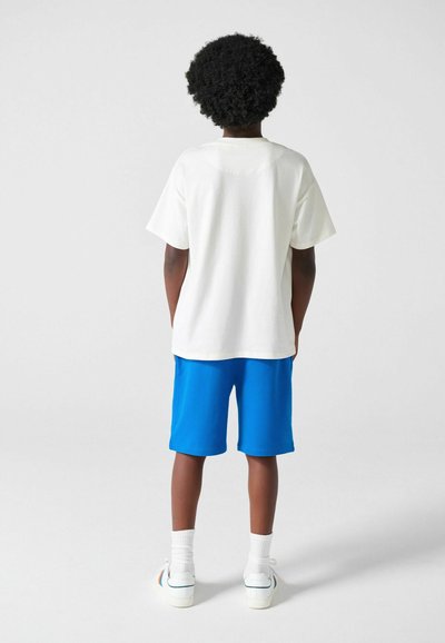 Niño con cabello rizado, vistiendo una camiseta blanca extragrande, pantalones cortos azul brillante, calcetas blancas y zapatillas blancas, de espaldas sobre un fondo liso.