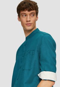 Jonge man in een teal overhemd met opgerolde mouwen, die zijwaarts kijkt tegen een effen lichtgrijze achtergrond.
