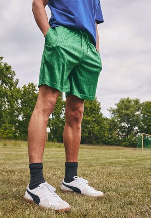 Green athletic shorts with a textured pattern, drawstring waist, side pockets, paired with white and black sneakers and black socks.