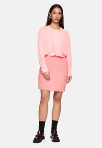 Rosa Langarmbluse mit Falten, kombiniert mit einem texturierten rosa Minirock. Model trägt schwarze Socken und glänzende burgunderfarbene Schuhe.