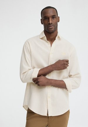 Hombre con el pelo corto que lleva una camisa beige clara abotonada y pantalones marrones claros, ajustándose una manga remangada, sobre un fondo blanco liso.