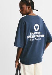 Marineblå oversized t-shirt med korte ærmer, der har hvid tekst: "DARE MORE WAKE MEMORIES" og et hjerte symbol på ryggen.