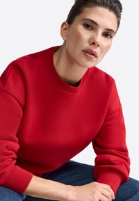 Červená mikina s okrúhlym výstrihom a rebrovanými manžetami. Mäkká textúra s voľným strihom, spárovaná s modrými džínsami.