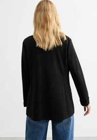 Blazer negro con un diseño de corte recto, mangas largas con puños enrollados y una textura suave. Llevado sobre unos jeans azules, visto desde la parte posterior.