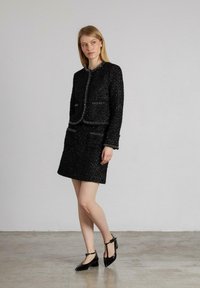 Schwarze Tweedjacke mit taillierter Silhouette, strukturiertem Stoff und zwei vorderen Taschen, kombiniert mit einem passenden Minirock und schwarzen Lackpumps.