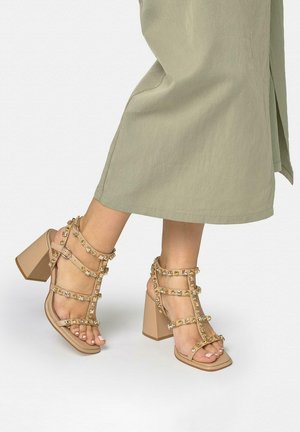 Pies con sandalias de tacón alto beige con tachuelas doradas y tiras alrededor del tobillo, combinadas con pantalones anchos de pierna verde claro recortados por encima de los tobillos.