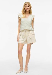 Lys grøn strikket bluse med V-hals og flæseærmer, parret med beige shorts med snoretræk, som har sidelommer og en blød tekstur.