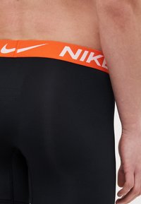 Fekete sportnadrág narancs derékrésszel, rajta fehér "NIKE" felirattal, sima textúrájú, testhez simuló fazon. A varrás mentén látható a steppelés.