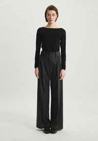 Haut noir à manches longues et pantalon à taille haute, à largas rayures. Le pantalon possède des plis et est fabriqué dans un tissu texturé. Chaussures sombres.