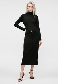 Žena s dlhými blond vlasmi, oblečená v čiernych midi šatách s dlhým rukávom, so zlatou prackou na opasku a čiernymi lodičkami na členkovom pásku, stojí proti bielej stene.