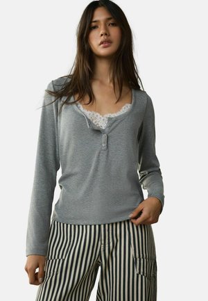 Nő szürke hosszú ujjú henley ingben, fehér csipke felsővel, fekete-fehér, magas derekú, függőleges csíkos nadrággal párosítva.