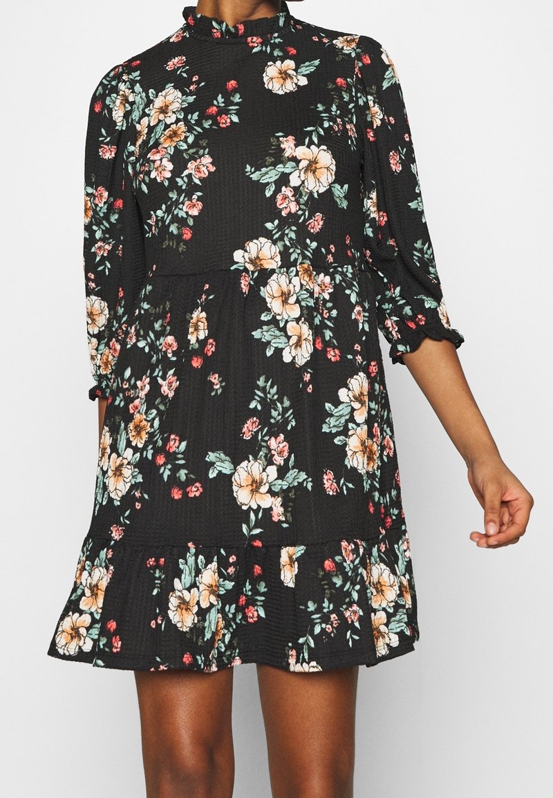 Schwarzes Blumenkleid mit langen Ärmeln und gerafftem Saum. Es hat pinke und gelbe Blumen, strukturierte Stoffe und einen hohen Kragen.