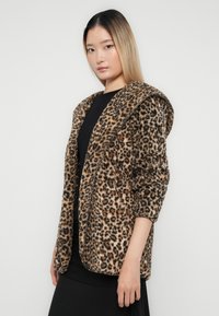 Mladá žena so rovnými blond vlasmi, oblečená v hnedo-čiernej bundě s leopardím vzorom, stojí proti bielej stene a má na sebe čierny outfit.