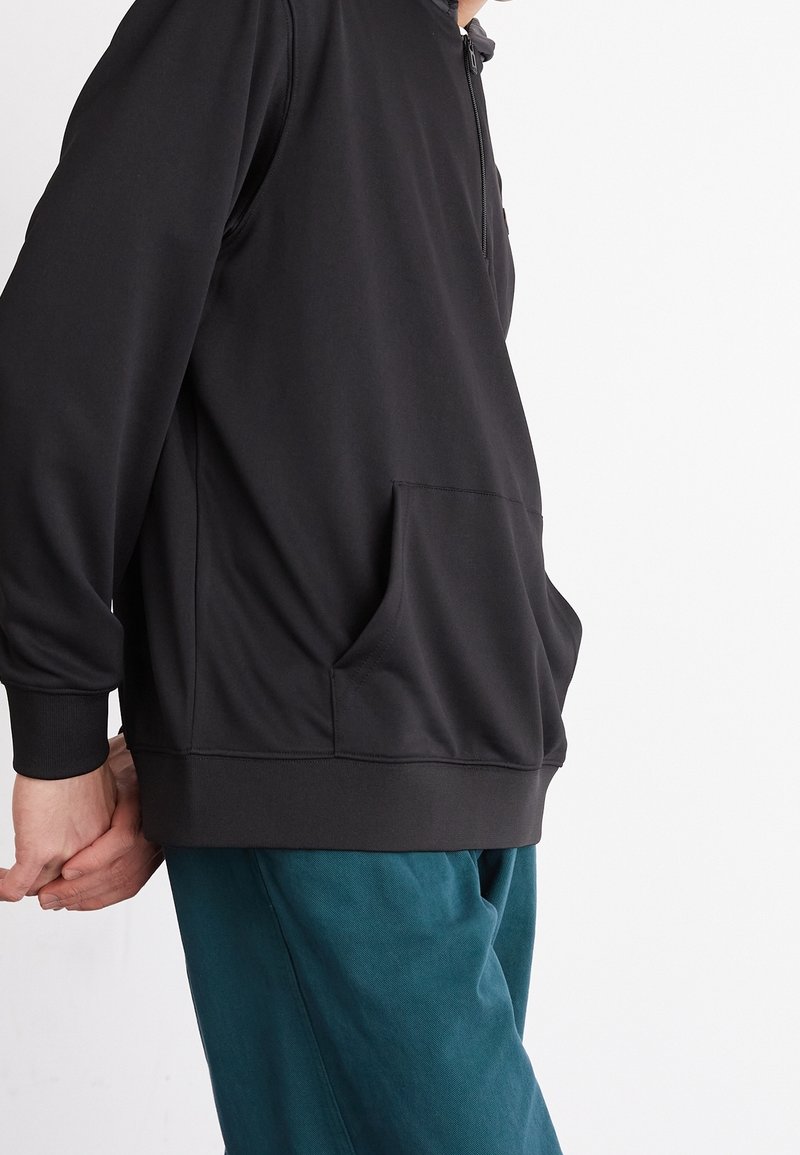 Felpa nera con cappuccio realizzata in morbido tessuto, dotata di tasca frontale e polsini a coste, abbinata a pantaloni teal. Texture liscia e design minimalista.