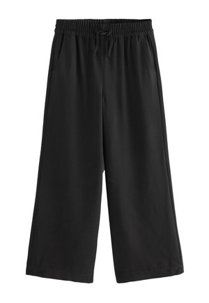 Mädchen steht vor einer beigen Wand und trägt ein weißes Poloshirt mit kurzen Ärmeln, schwarze weite Hosen und schwarze Slipper.