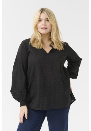 Blouse noire avec un décolleté en V et un motif à pois subtil, dotée de longues manches bouffantes avec des poignets élastiques et une coupe décontractée. Associée à un jean foncé.