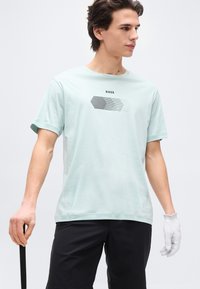 Νεαρός άνδρας με ανοιχτό μπλε γραφικό t-shirt BOSS, μαύρο σορτς και λευκό γάντι γκολφ, κρατώντας μια μαύρη ράβδο γκολφ.