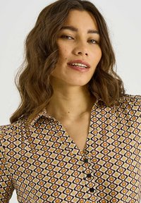 Blusa de manga larga con un patrón geométrico en colores cálidos. Presenta un cuello abotonado y una tela texturizada con acentos en marrón, amarillo y gris.