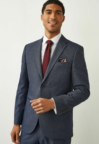 Veste de costume bleu marine à motif avec revers crantés, accents de boutons marron et chemise blanche. Une cravate bordeaux et un mouchoir de poche complètent le look.