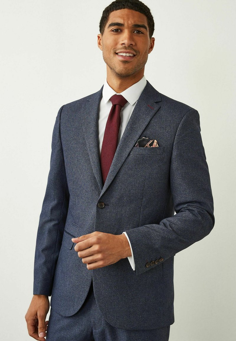 Veste de costume bleu marine à motif avec revers crantés, accents de boutons marron et chemise blanche. Une cravate bordeaux et un mouchoir de poche complètent le look.