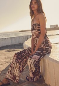 Kombinezon bez naramenica u smešnim smeškim i ružičastim uzorcima, sa širokim nogavicama. Model sjedi na kamenoj ivici s pozadinom plaže, noseći crne sandale.