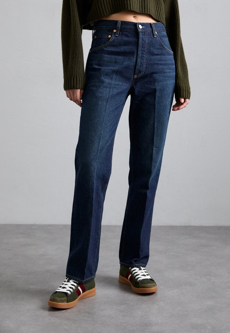 Dunkelblaue Jeans im geraden Schnitt, ausgestattet mit fünf Taschen, Gürtelschlaufen und einer Knopfleiste. Kombiniert mit grünen Sneakers mit weißen Akzenten.