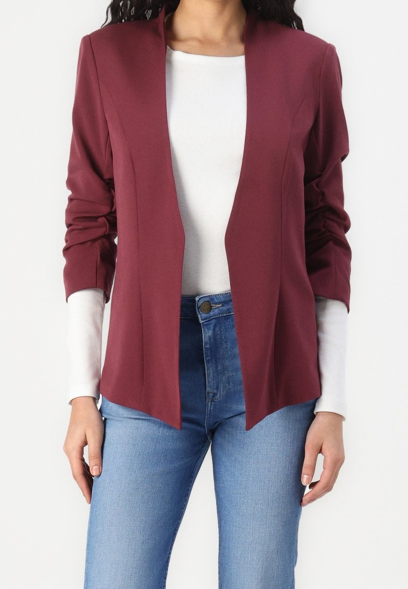 Bordeauxrode blazer met sjaalkraag, getailleerde pasvorm, lange mouwen en een open voorkant, gedragen over een wit overhemd met lange mouwen en blauwe spijkerbroek.