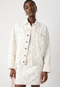 Veste en denim blanche avec des motifs floraux roses, boutons en argent et deux poches avant, associée à une jupe en denim assortie.